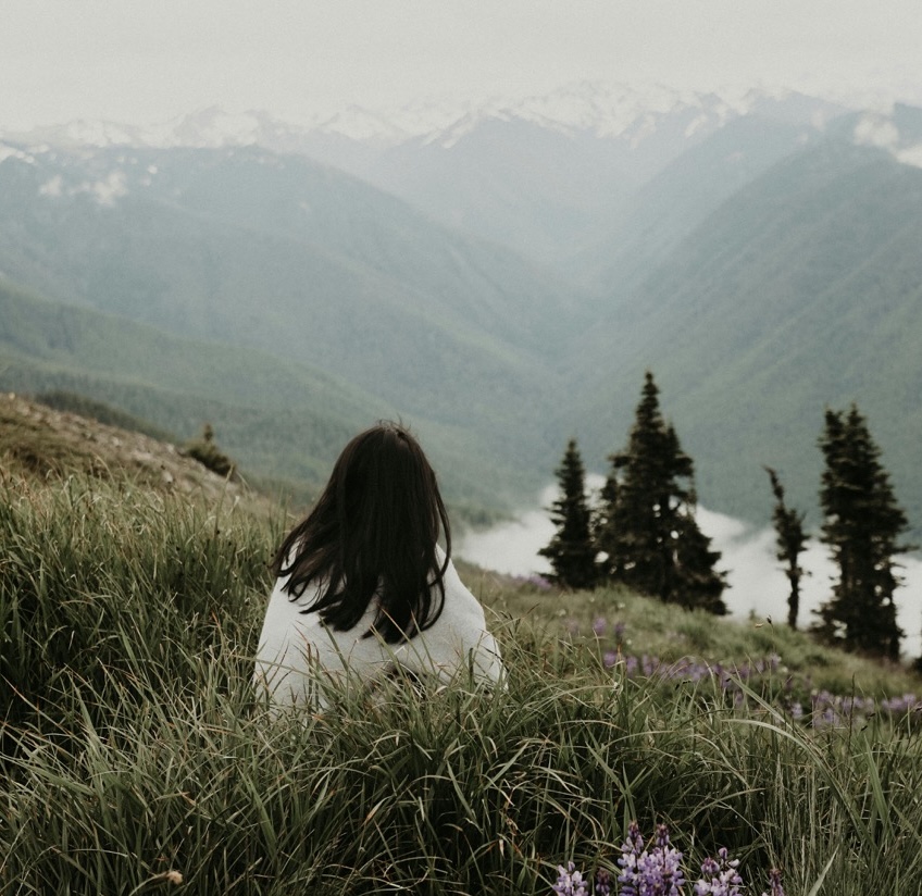 Femme assise dans l'herbe, de dos, regardant un paysage de montagnes